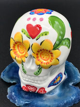 Load image into Gallery viewer, Inspired by nature, this ceramic skull statuette is full of vibrant color and intricate details. The primary color is white with multi-color floral motifs that are painted across the skull, along with hearts and leaves and lively colorful accents around the entire skull.

