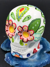 Load image into Gallery viewer, Inspired by nature, this ceramic skull statuette is full of vibrant color and intricate details that I hand painted. The primary color is white with multi-color floral motifs that are painted across the skull, along with hearts and leaves and lively colorful accents around the entire skull. 
