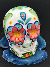 Load image into Gallery viewer, Inspired by nature, this ceramic skull statuette is full of vibrant color and intricate details. The primary color is white with multi-color floral motifs that are painted across the 
