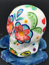 Load image into Gallery viewer, Inspired by nature, this ceramic skull statuette is full of vibrant color and intricate details. The primary color is white with multi-color floral motifs that are painted across the 

