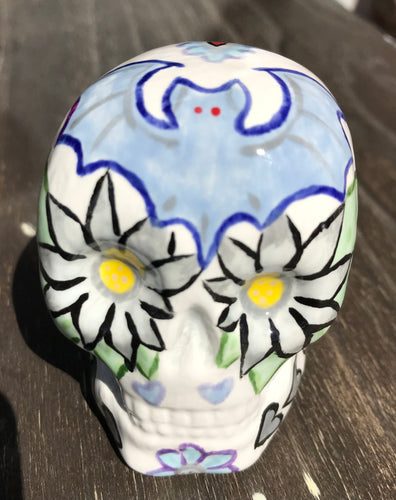 This ceramic skull art is a unique and eye-catching combination of bright colors and dark tones. It features two bat silhouettes and flowers in warm colors like red, yellow, and green, as well as medium gray and royal blue. Its vibrant red eyes give the piece an inviting touch. Perfect for a mini goth witchy altar, this figurine is sure to stand out. Measurements: H 2 3/4” x W 2” x D 2”