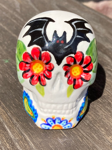 This Bat Lovers Ceramic Skull Art is a unique addition to any dark gothic collection. Featuring a mini batty with bright red eyes and outlined in royal blue, accented with white lines to highlight its small bones and an additional bat with vibrant red eyes on the opposite side. Enjoy the playful contrast of warm tones and a darker theme in this eye-catching figurine. Measurements: H 2 3/4” x W 2” x D 2”