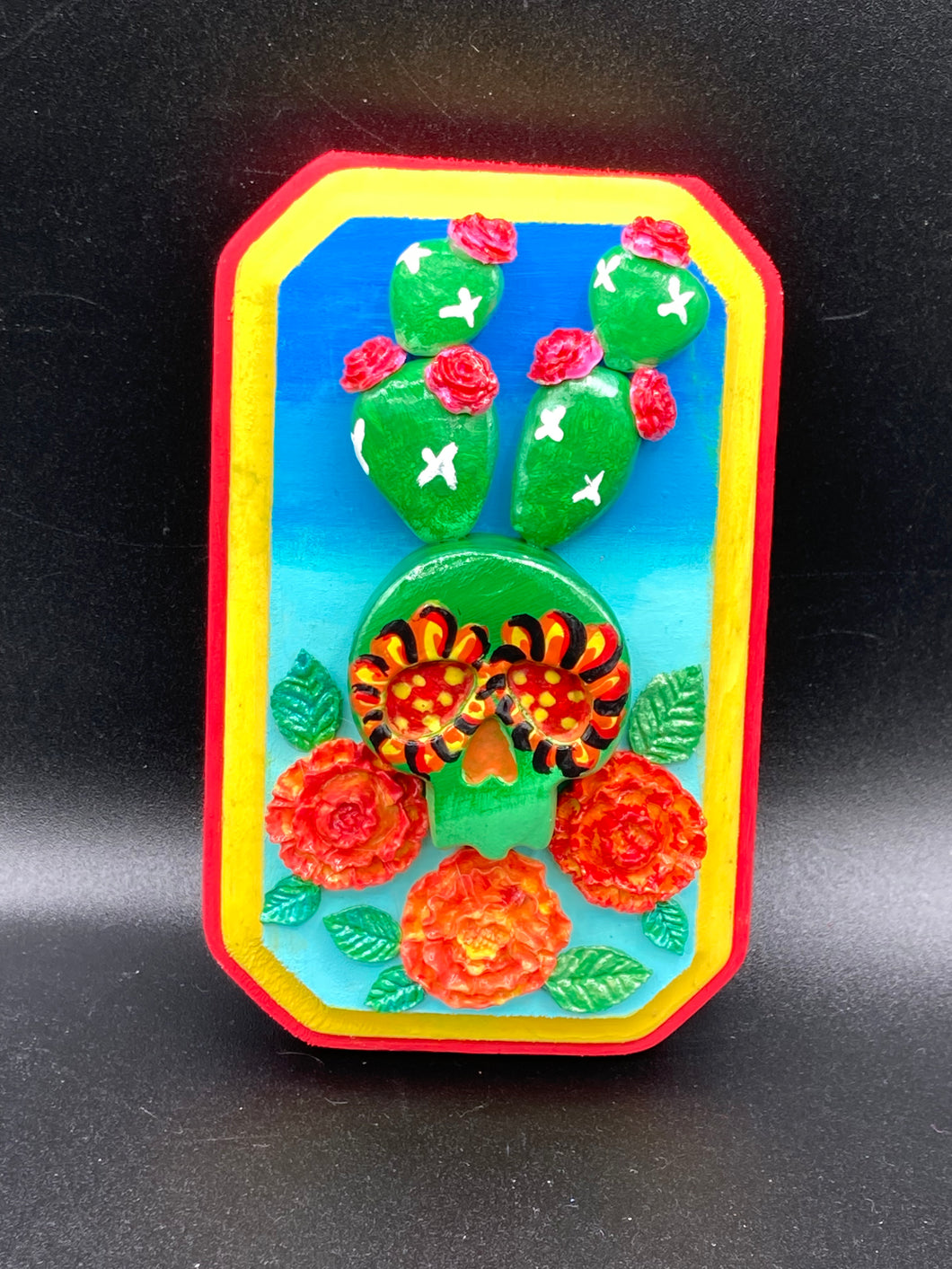 This handcrafted polymer clay wall art features a wood frame painted in a gradient from light to dark blue, creating a stunning starry night sky look. With a bright green skull cactus plant and bright yellow blooming flowers. Boasting vivid marigold blooms and a bright yellow and red hue frame to make a bold contrast with the warm background hue. This piece is sure to make a statement.