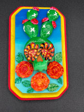 Load image into Gallery viewer, This handcrafted polymer clay wall art features a wood frame painted in a gradient from light to dark blue, creating a stunning starry night sky look. With a bright green skull cactus plant and bright yellow blooming flowers. Boasting vivid marigold blooms and a bright yellow and red hue frame to make a bold contrast with the warm background hue. This piece is sure to make a statement.
