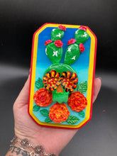 Load image into Gallery viewer, This handcrafted polymer clay wall art features a wood frame painted in a gradient from light to dark blue, creating a stunning starry night sky look. With a bright green skull cactus plant and bright yellow blooming flowers. Boasting vivid marigold blooms and a bright yellow and red hue frame to make a bold contrast with the warm background hue. This piece is sure to make a statement.
