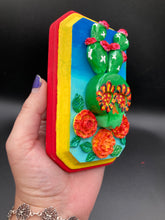 Load image into Gallery viewer, This handcrafted polymer clay wall art features a wood frame painted in a gradient from light to dark blue, creating a stunning starry night sky look. With a bright green skull cactus plant and bright yellow blooming flowers. Boasting vivid marigold blooms and a bright yellow and red hue frame to make a bold contrast with the warm background hue. This piece is sure to make a statement.

