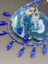 Load image into Gallery viewer, This set of large, eye-catching chandelier-style earrings draw inspiration from the vibrant array of colors found in the galaxy and cosmos. Featuring three strands of shimmering glass beads in bold shades of blue, purple, gold, and green, it's an essential piece for any jewelry collection.  Measurements: 3 1/2” in length
