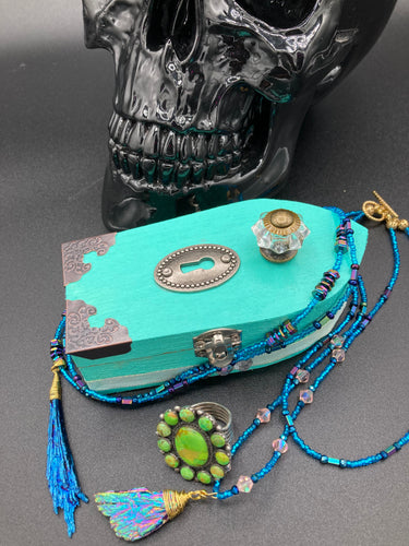 This Whimsical Fairy Wood Coffin Box is a truly unique and magical piece of home decor. With its vibrant turquoise exterior and eye-catching mini door knob, this Steampunk-inspired box is sure to be a conversation starter. Inside, the glittery gold interior and colorful butterfly detailing will bring a touch of fantasy to any room. The silver key and accompanying iridescent crystals complete the mystical and pastel goth look!