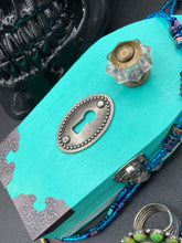 Load image into Gallery viewer, This Whimsical Fairy Wood Coffin Box is a truly unique and magical piece of home decor. With its vibrant turquoise exterior and eye-catching mini door knob, this Steampunk-inspired box is sure to be a conversation starter. Inside, the glittery gold interior and colorful butterfly detailing will bring a touch of fantasy to any room. The silver key and accompanying iridescent crystals complete the mystical and pastel goth look!
