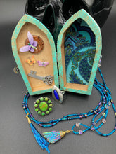 Load image into Gallery viewer, This Whimsical Fairy Wood Coffin Box is a truly unique and magical piece of home decor. With its vibrant turquoise exterior and eye-catching mini door knob, this Steampunk-inspired box is sure to be a conversation starter. Inside, the glittery gold interior and colorful butterfly detailing will bring a touch of fantasy to any room. The silver key and accompanying iridescent crystals complete the mystical and pastel goth look!
