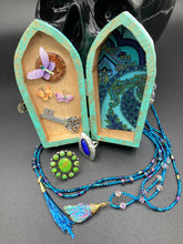Load image into Gallery viewer, This Whimsical Fairy Wood Coffin Box is a truly unique and magical piece of home decor. With its vibrant turquoise exterior and eye-catching mini door knob, this Steampunk-inspired box is sure to be a conversation starter. Inside, the glittery gold interior and colorful butterfly detailing will bring a touch of fantasy to any room. The silver key and accompanying iridescent crystals complete the mystical and pastel goth look!
