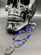 Load image into Gallery viewer, This spooky resin anatomical skeleton hand pendant is a perfect handy jewelry piece! The anatomical skeleton hand is made of resin with brass metal backing. Rich royal blue glass beads to give it a huge pop of color. Rectangular and hexagon rainbow hematite beads to compliment the shapes of the finger bones in the pendant. Bright canary yellow beads along the strands to break up the cool hues a bit. Necklace measures 16” adjustable up to 18” in length, pendant is 1 1/4”
