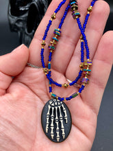 Load image into Gallery viewer, This spooky resin anatomical skeleton hand pendant is a perfect handy jewelry piece! The anatomical skeleton hand is made of resin with brass metal backing. Rich royal blue glass beads to give it a huge pop of color. Rectangular and hexagon rainbow hematite beads to compliment the shapes of the finger bones in the pendant. Bright canary yellow beads along the strands to break up the cool hues a bit. Necklace measures 16” adjustable up to 18” in length, pendant is 1 1/4”
