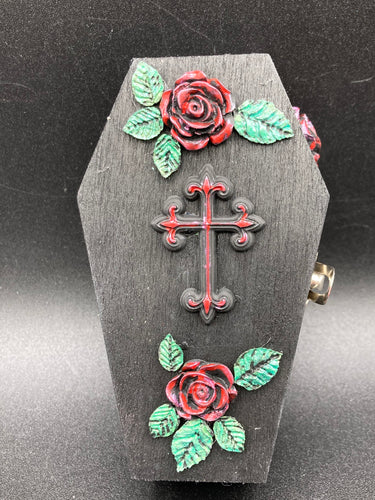 Keep your special jewelry safe in this vintage-inspired wooden coffin jewelry box. Crafted with attention to detail, the box features a black and red cross design, surrounded by dark red roses and a black glittery ribbon. The interior is adorned with a black and silver cross and a bright red crystal in the center. A stylish and secure way to store your beloved accessories.