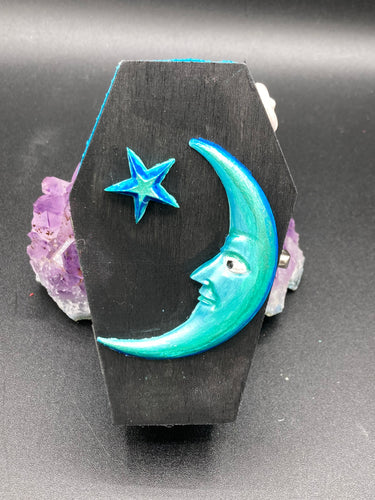 This sleek black box has an eye-catching turquoise crescent at its center, accompanied by a bright blue ribbon and a dainty white moon. Inside, the interior is decorated with a vivid royal blue and detailed with a small crescent moon and black-and-silver cross.  Coffin Dimensions: Length: 4 1/2 Inches; Width: 2 1/2 inches; Height: 1 5/8 Inches