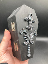 Load image into Gallery viewer, This vintage-inspired coffin ring box will make a spooky yet cute addition to any jewelry collection. The sleek black box has eye-catching black and gray embellishments, while the interior is beautifully decorated with a vivid turquoise blue fabric and a highly detailed black and silver cross with dark red roses. Function and style meet with this unique and stylish piece.
