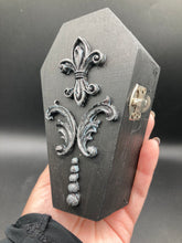 Load image into Gallery viewer, This vintage-inspired coffin ring box will make a spooky yet cute addition to any jewelry collection. The sleek black box has eye-catching black and gray embellishments, while the interior is beautifully decorated with a vivid turquoise blue fabric and a highly detailed black and silver cross with dark red roses. Function and style meet with this unique and stylish piece.
