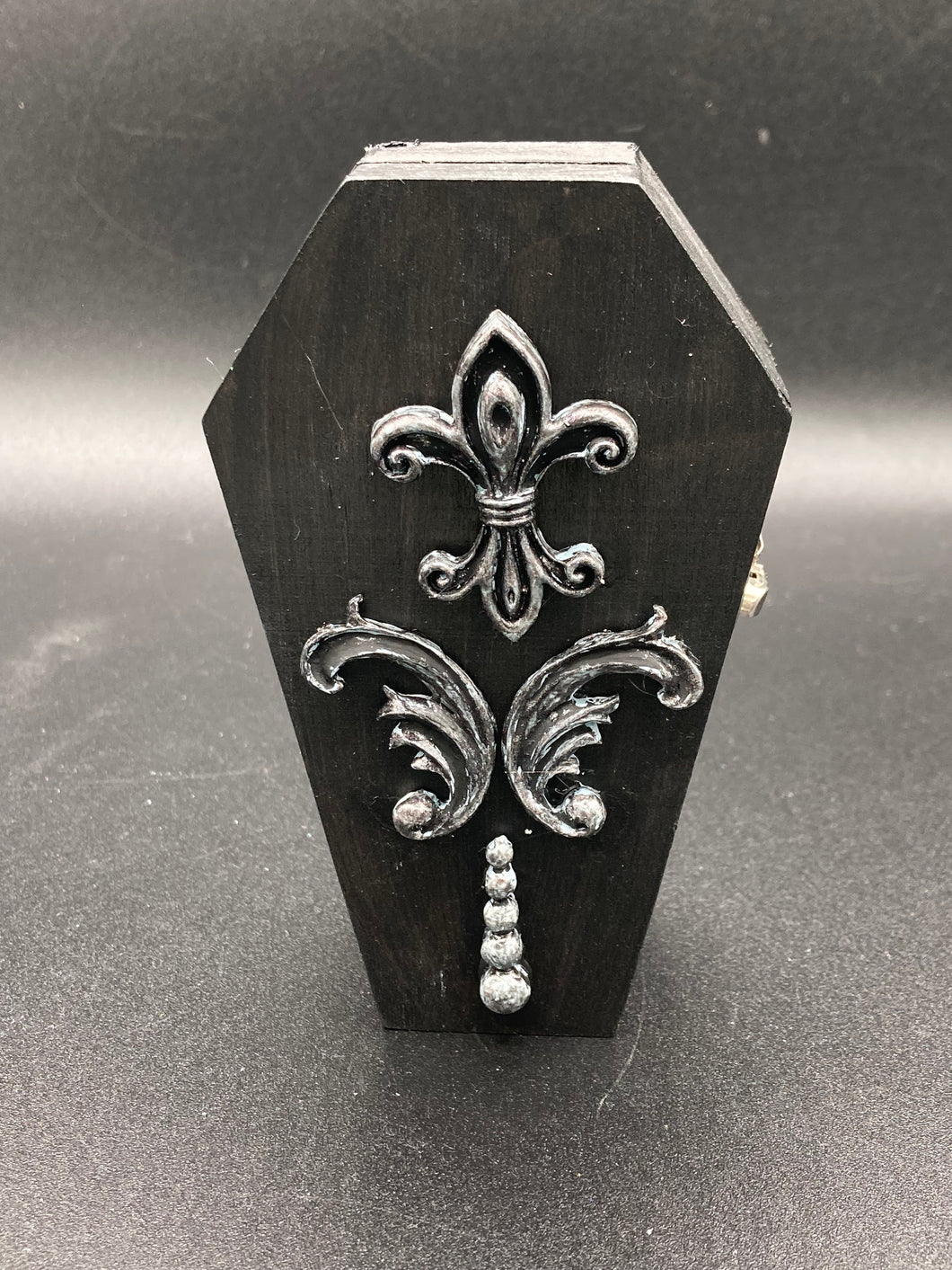 This vintage-inspired coffin ring box will make a spooky yet cute addition to any jewelry collection. The sleek black box has eye-catching black and gray embellishments, while the interior is beautifully decorated with a vivid turquoise blue fabric and a highly detailed black and silver cross with dark red roses. Function and style meet with this unique and stylish piece.