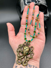 Load image into Gallery viewer, This Steampunk Brass Octopus Emerald Eyes Choker Style Necklace is an eye-catching piece for cosplay and gothic aficionados alike. Crafted with a large brass octopus pendant, emerald green eyes, and an adjustable glass beaded strand, this necklace is sure to turn heads. Featuring a range of light and dark green, gold, and brass tones, this necklace complements any sci-fi, fantasy, or steampunk ensemble.  Measurements: 15 1/2” adjustable up to 17" in length
