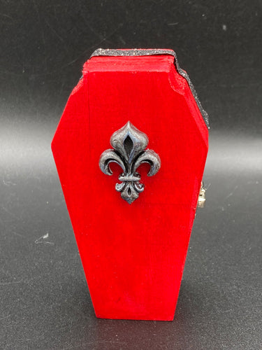 Super vibrant red coffin box has an eye-catching black and gray fleur de lis embellishment in the  center, accompanied by a glittery black ribbon around the coffin. Inside, the interior is all black with a floral detailed black-silver cross.