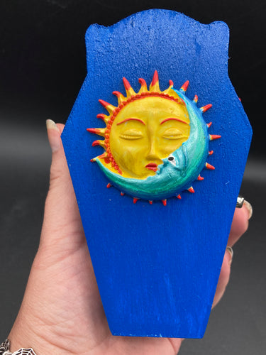 This coffin-shaped box features a bright royal blue exterior, with a bold sun and crescent adorning the center. Complemented with a shimmery pastel-yellow ribbon and flower, the inside is painted in a canary yellow hue with turquoise crescent moon detailing and traditional Mexican-themed fabric.  Coffin Dimensions: Length: 4 1/2 Inches; Width: 2 1/2 inches; Height: 1 5/8 Inches