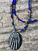 Load image into Gallery viewer, This spooky resin anatomical skeleton hand pendant is a perfect handy jewelry piece! The anatomical skeleton hand is made of resin with brass metal backing. Rich royal blue glass beads to give it a huge pop of color. Rectangular and hexagon rainbow hematite beads to compliment the shapes of the finger bones in the pendant. Bright canary yellow beads along the strands to break up the cool hues a bit. Necklace measures 16” adjustable up to 18” in length, pendant is 1 1/4”
