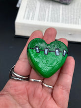 Load image into Gallery viewer, Perfect cute and spooky Halloween item to add to your fridge, locker, cubicle or toolbox. This hand-crafted light and dark green heart-shaped magnet showcases a Frankenstein-inspired design.  Measurements: H 1 1/2” x W 1 5/8” x D 1/2”
