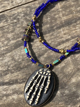 Load image into Gallery viewer, This spooky resin anatomical skeleton hand pendant is a perfect handy jewelry piece! The anatomical skeleton hand is made of resin with brass metal backing. Rich royal blue glass beads to give it a huge pop of color. Rectangular and hexagon rainbow hematite beads to compliment the shapes of the finger bones in the pendant. Bright canary yellow beads along the strands to break up the cool hues a bit. Necklace measures 16” adjustable up to 18” in length, pendant is 1 1/4”
