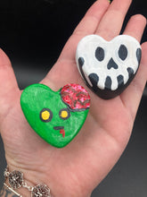 Load image into Gallery viewer, Perfect cute and spooky Halloween magnet art to add to your fridge, locker, cubicle or toolbox.  This hand-crafted black heart-shaped magnet features a white skull figurine  with distinctive black eyes. Cute heart-shaped green zombie with exposed brains magnet.  Measurements: H 1 1/2” x W 1 5/8” x D 1/2”
