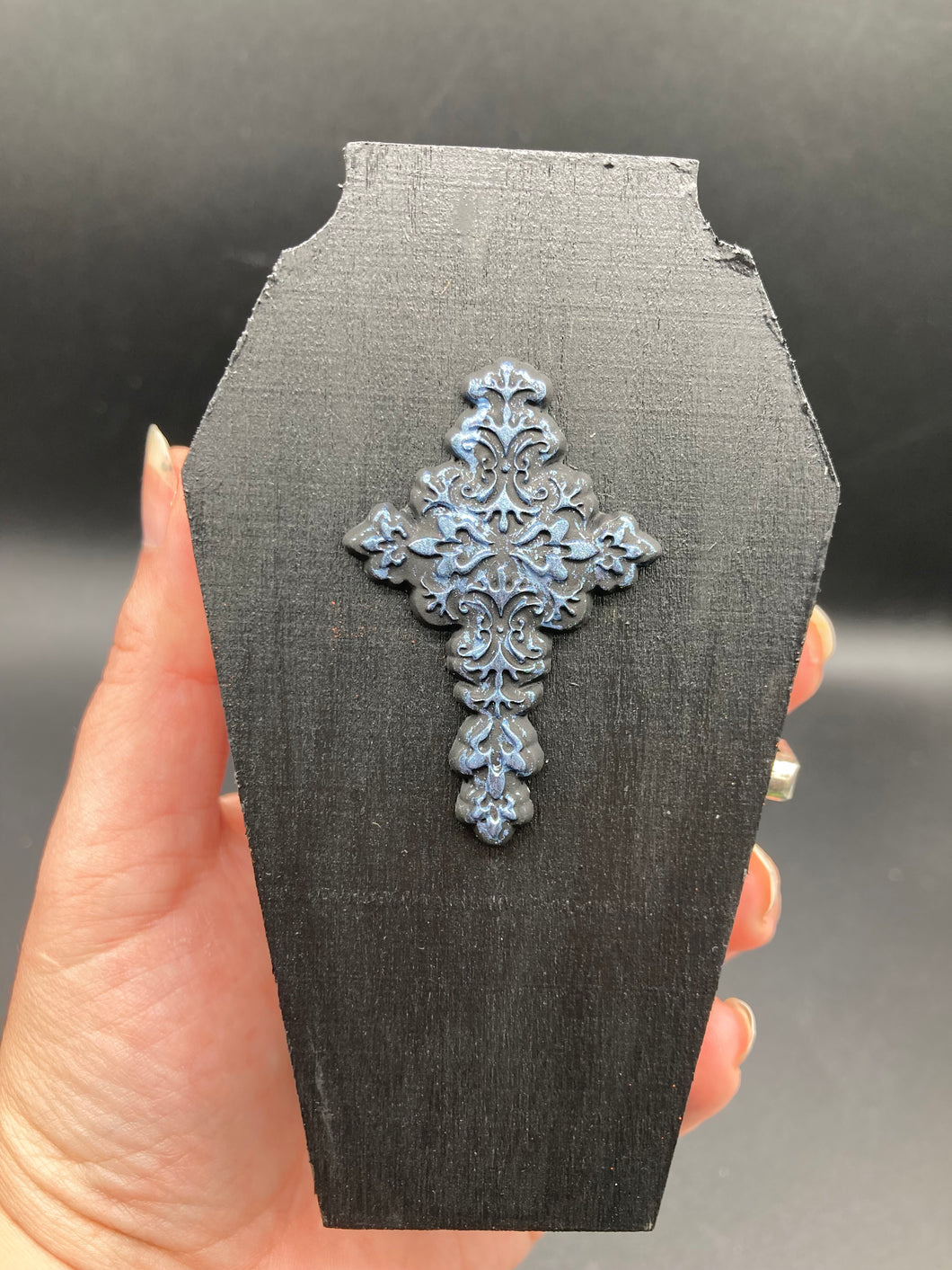 This unique vintage-inspired ring box is perfect for storing your jewelry and other special items. The wood coffin design is accented with a detailed floral black silver cross and a white and black ribbon. The interior is lined with a skull fabric and a fleur de lis embellishment, adding a spooky yet cute flair.