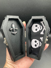 Load image into Gallery viewer, This unique vintage-inspired ring box is perfect for storing your jewelry and other special items. The wood coffin design is accented with a detailed floral black silver cross and a white and black ribbon. The interior is lined with a skull fabric and a fleur de lis embellishment, adding a spooky yet cute flair.
