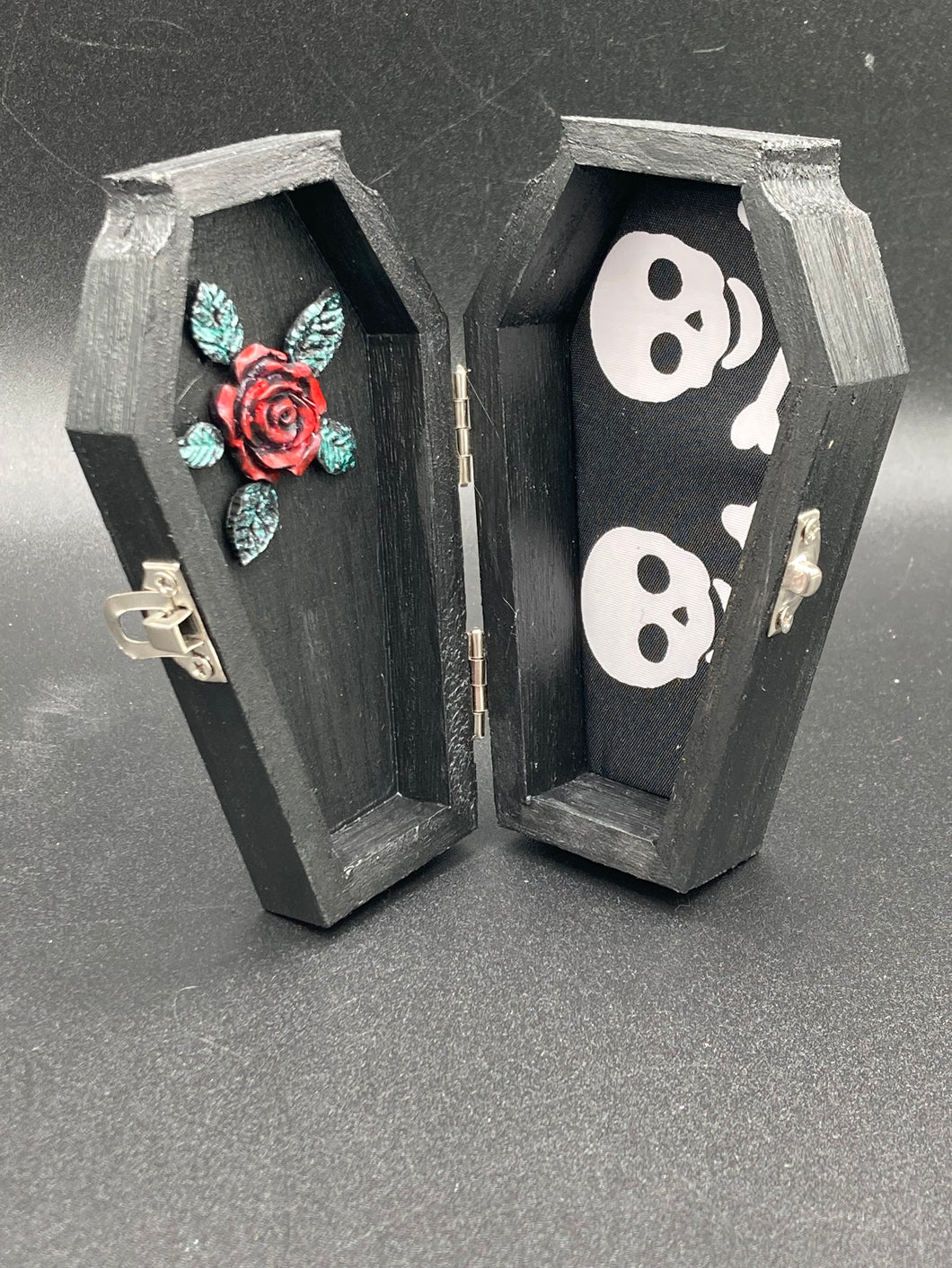 This unique vintage-inspired ring box is perfect for storing your jewelry and other special items. The wood coffin design is accented with a detailed black silver cross. The interior is lined with a skull fabric and a large dark red rose, adding a spooky yet elegant flair.