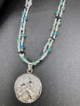 Load image into Gallery viewer, This stylish necklace features a shimmery sterling silver quartz pendant with cats and stars, making it the perfect accessory for any cat lover. With two-hole silver and black beads that stand out against the light aquamarine glass beads, this double strand necklace holds a round kitty pendant made with various hues of clear and iridescent glass beads. Add a touch of purrfection to your look with this statement piece.  Measurements:  Necklace is 15”adjustable up to 18&quot; 
