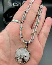 Load image into Gallery viewer, This stylish necklace features a shimmery sterling silver quartz pendant with cats and stars, making it the perfect accessory for any cat lover. With two-hole silver and black beads that stand out against the light aquamarine glass beads, this double strand necklace holds a round kitty pendant made with various hues of clear and iridescent glass beads. Add a touch of purrfection to your look with this statement piece.  Measurements:  Necklace is 15”adjustable up to 18&quot; 
