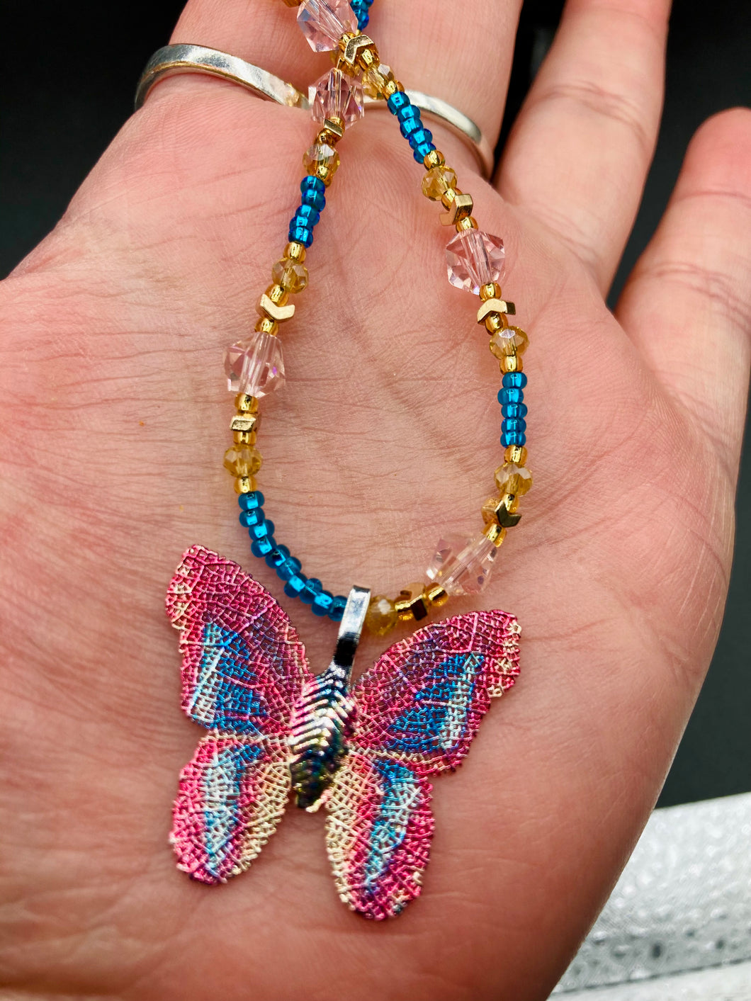 This delicate butterfly pendant is crafted from a basil leaf and adorned with accents of pink, gold and blue. A shining glass beaded necklace crafted from beads of varying shades of pink, gold, and blue, creates a beautiful accompaniment to the butterfly wings. Wear this ravishing necklace and butterfly jewelry pendant to embody the spirit of warm Spring and Summer days. 16 1/4” in length adjustable up to 18