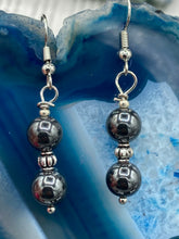 Load image into Gallery viewer, Elevate your boho chic style with these beautiful hematite and glass beaded earrings. Featuring two hematite round beads and a floral silver metal bead in the middle, these earrings offer a stylish and chic touch to any outfit.  Measurements: 1 1/4” in length 
