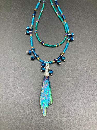 This celestial and galaxy goddess necklace features two strands of glass beading and a captivating Kyanite wired stone pendant. Its mixture of shimmering purples, blues, greens, and golds is truly striking and provides an alluring look like no other. Add this bold and beautiful jewelry piece to your collection today for a statement look that truly stands out. Necklace is 15” in length, can be adjusted to 18