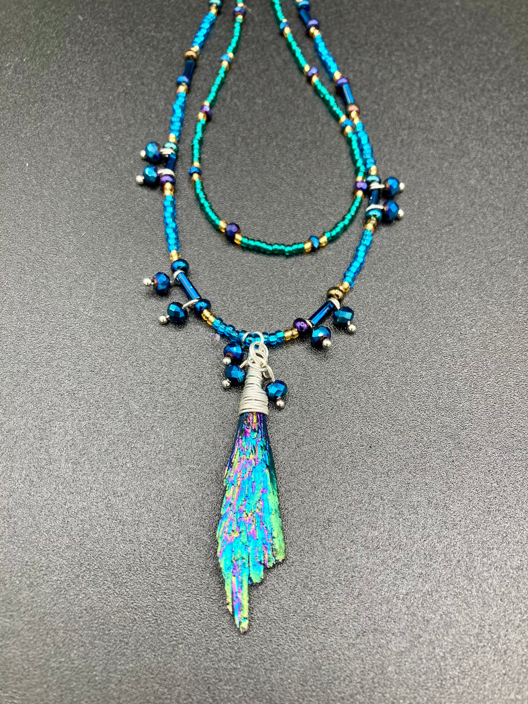 This celestial and galaxy goddess necklace features two strands of glass beading and a captivating Kyanite wired stone pendant. Its mixture of shimmering purples, blues, greens, and golds is truly striking and provides an alluring look like no other. Add this bold and beautiful jewelry piece to your collection today for a statement look that truly stands out. Necklace is 15” in length, can be adjusted to 18