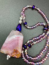 Load image into Gallery viewer, A soft dusty rose agate pendant is accompanied by shimmery beads of dusty rose, blue, dark pink, and gold. Adorned with large deep purple glass beads with a hint of gold shimmer, this stunning necklace set evokes a rose garden.  Measurements:  Necklace is 15” adjustable to 18&quot;
