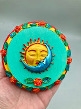 Load image into Gallery viewer, Handcrafted polymer clay wall art with a wood frame, featuring a sun and moon motif complemented with contrasting cool and warm colors. The background is a bright turquoise hue, with marigold flowers and leaves adorning the frame.
