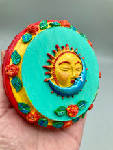 Load image into Gallery viewer, Handcrafted polymer clay wall art with a wood frame, featuring a sun and moon motif complemented with contrasting cool and warm colors. The background is a bright turquoise hue, with marigold flowers and leaves adorning the frame.
