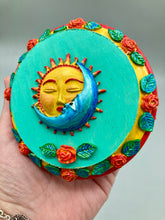 Load image into Gallery viewer, Handcrafted polymer clay wall art with a wood frame, featuring a sun and moon motif complemented with contrasting cool and warm colors. The background is a bright turquoise hue, with marigold flowers and leaves adorning the frame.
