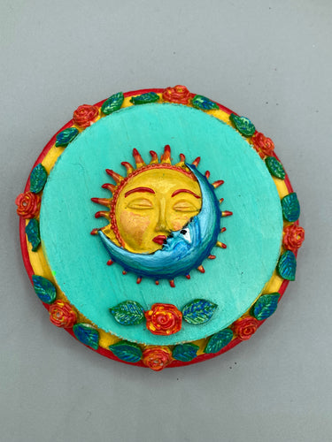 Handcrafted polymer clay wall art with a wood frame, featuring a sun and moon motif complemented with contrasting cool and warm colors. The background is a bright turquoise hue, with marigold flowers and leaves adorning the frame.