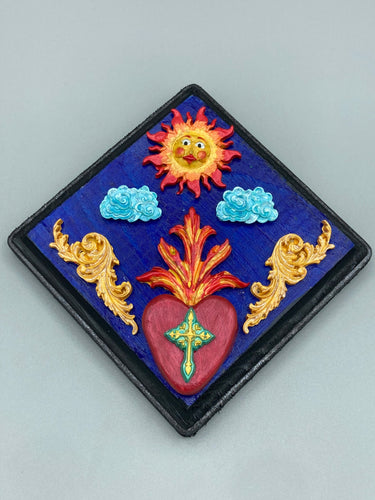 This handcrafted polymer clay wall art is displayed on a square wood frame, featuring sun and light blue cloud motifs on each side and a dark royal blue backdrop with a bright red sacred heart in the center. A turquoise and yellow cross provides contrast to the warm toned heart, which is further embellished with gold accents on each side of the heart. H 6 3/4