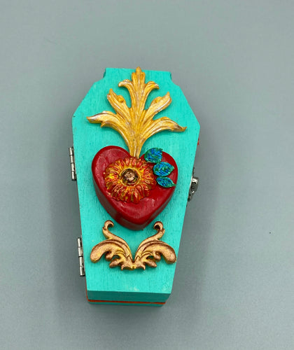 This coffin-shaped box features a bright turquoise exterior, with a hand-crafted bright red heart. The heart is detailed with a sunflower and vibrant green leaves, with a vibrant gold fire embellishment over the center. Gold decorative embellishments at the bottom of the heart add a luxurious touch. Complemented with a shimmery bright orange ribbon and flower around the coffin box, the inside is painted in a vibrant orange hue with black and silver cross and traditional Mexican-themed fabric.