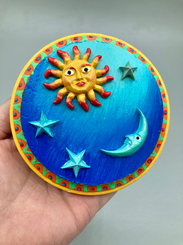 This handcrafted polymer clay wall art on a circle-shaped wood frame with a sun and crescent moon motifs on opposite side and stars. Its cool sky hues, including bright turquoise and dark royal blue, are further complemented by the bright orange and yellow frame adds an extra layer of sophistication to the warm-toned artwork.  One of a kind art pieces come ready to hang with a sawtooth hangers in the back. Measurement 4