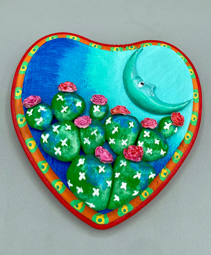 This handcrafted polymer clay wall art features a heart-shaped wood frame complimented with a crescent moon motif. The complimentary color palette consists of cool sky hues, from a bright turquoise to a dark royal blue. The piece is further enhanced with hand sculpted cacti plants with blooming pink flowers. The vibrant red and orange frame complements the cool-toned artwork. Measurements: H 5 1/4