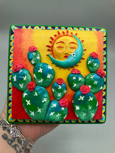 This handcrafted polymer clay wall art boasts a square-shaped wood frame with a sun and crescent moon motifs. Its warm sky hues, including yellow, orange and dark red, are further complemented by hand sculpted cacti plants with cool green-blue tones and dark pink flowers. The dark green frame adds an extra layer of sophistication to the warm-toned artwork.  One of a kind art pieces come ready to hang with a sawtooth hangers in the back. H 5