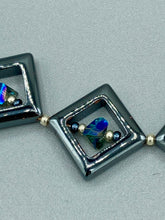 Load image into Gallery viewer, This two-strand necklace, crafted from aqua-blue and silver toned glass beads, is a stunning statement piece. The addition of faceted triangle-shaped glass beads tucked between hematite square beads adds an exquisite and unique flair. The necklace gracefully drapes around the neck, bringing an air of bold and elegant flair to any ensemble it complements. 15” in length, can be extended up to 17"
