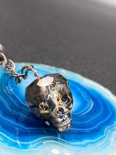 Load image into Gallery viewer, This beautiful edgy gun metal faceted shimmery multi-color glass skull bead keychain is full of hues of silver, purple, pink, blue, and gold. The split nickel key ring is 1 1/4&quot; in diameter and the entire key chain is 4 1/2&quot; in length (from key ring to skull bead)  Measurements: Nickel Free Key Ring is 1 1/4” in diameter, Entire Key Chain is 4 1/2&quot; in Length, Metal Skull Bead 5/8&quot; in Height
